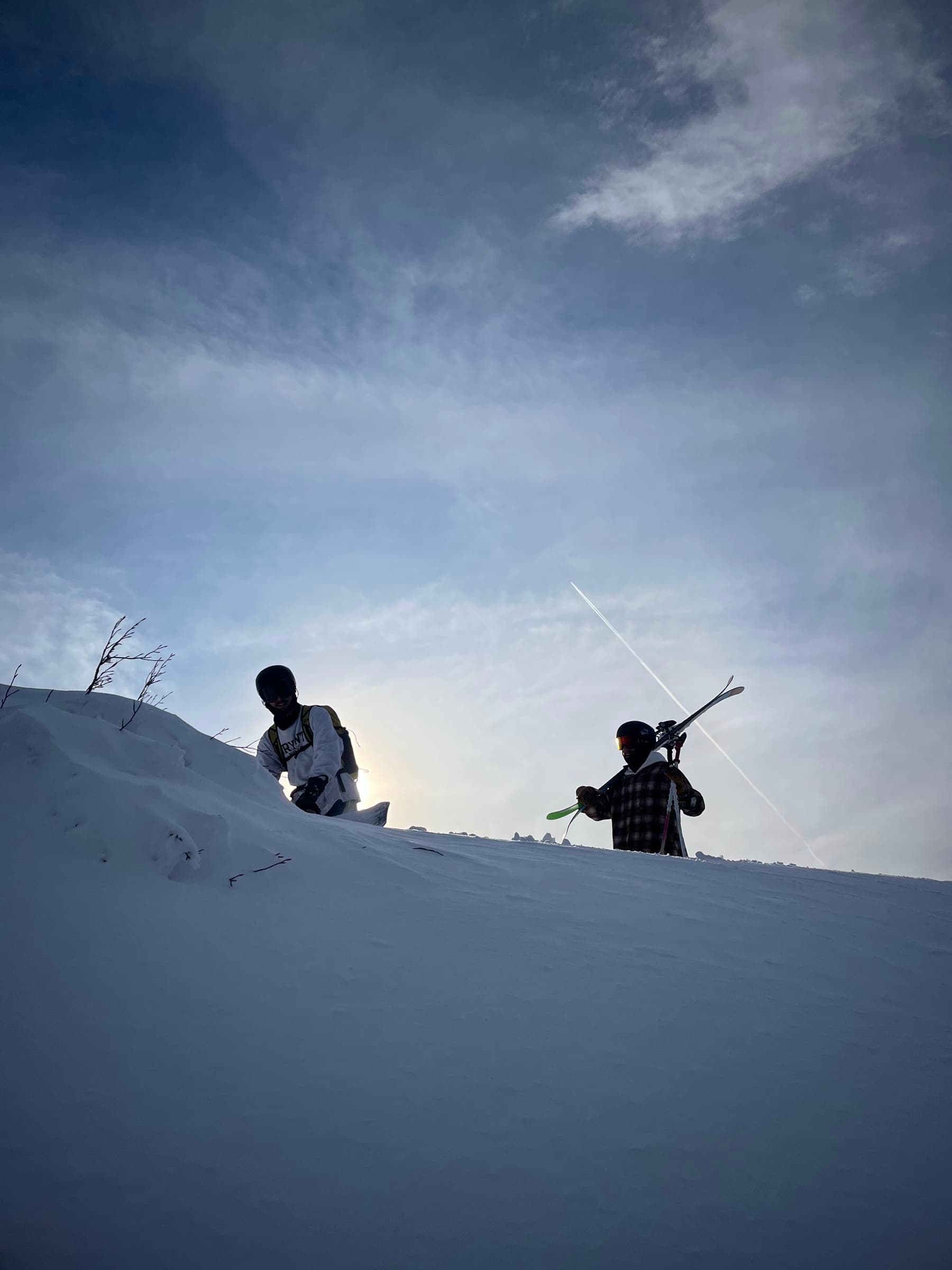 Two riders cresting a powder ridge at Madarao, skis over one shoulder, silhouetted against the winter sun.