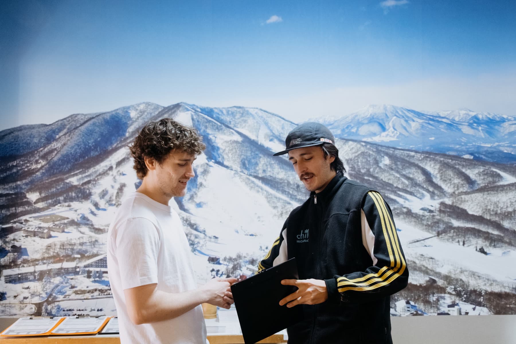 Two people talking over a snowboard outside the shop, the Madarao range behind them.